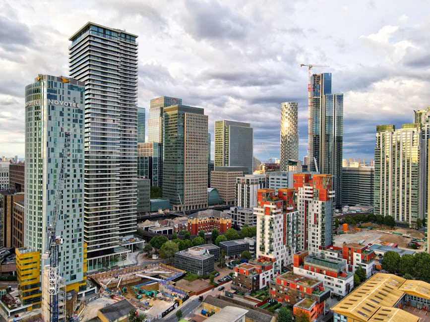 The image shows a vibrant cityscape of Canary Wharf in London, featuring a variety of modern skyscrapers with glass façades reflecting the sky and surrounding buildings. In the foreground, there are