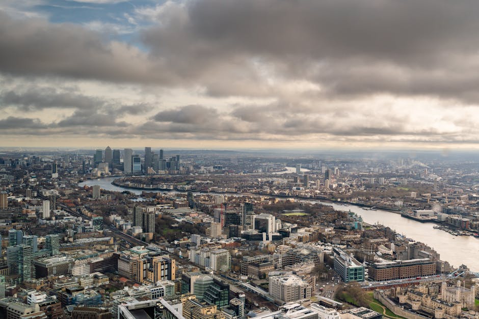 An aerial view of London showcasing a dense cityscape with numerous modern office buildings and residential structures, crossed by the winding River Thames. The scene features a cloudy sky overhead, c