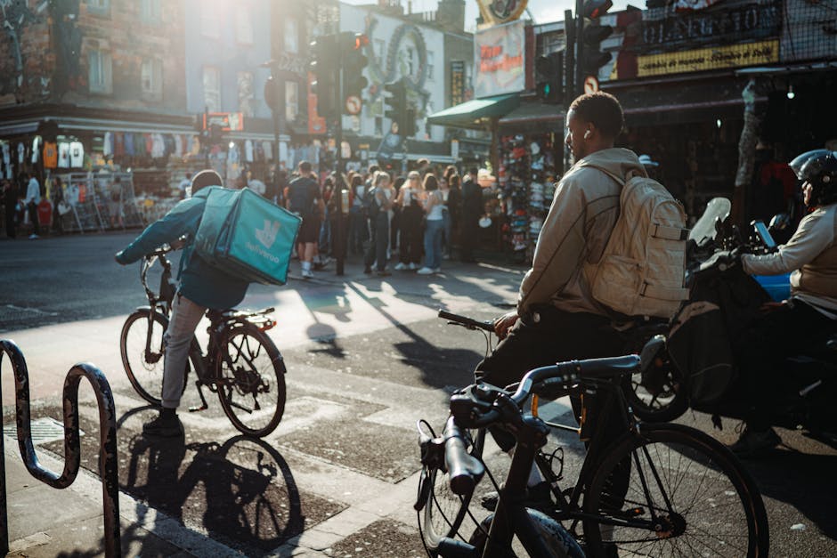 A busy street scene at Camden Market with two cyclists in the foreground; one cyclist wearing a beige jacket and carrying a light-colored backpack, sitting on an e-bike, while another cyclist in a blu