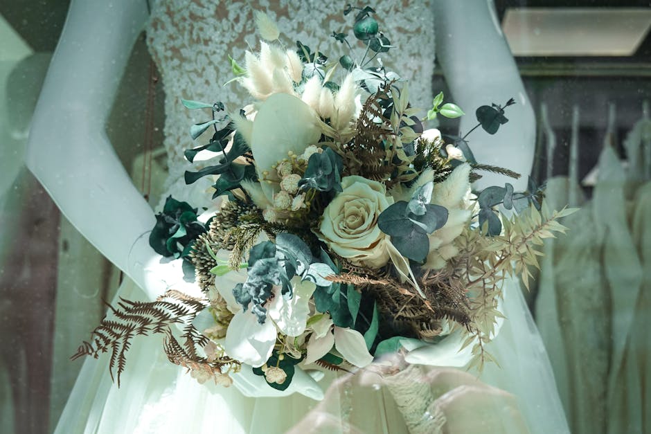 A bridal bouquet arrangement featuring cream and white roses, eucalyptus leaves, and dried ferns, with some soft, fluffy white accents. The bouquet is held by a person wearing a lace wedding dress, an