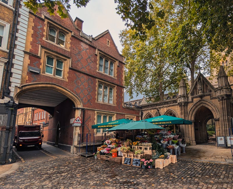 A vibrant flower stall set up under green umbrellas on a cobblestone street near a historic brick and stone archway in an urban area. The stall displays a colorful variety of fresh flowers, including 