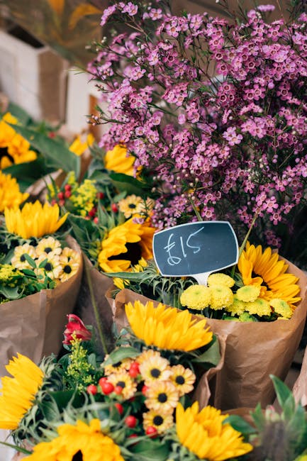 A vibrant display of fresh floral arrangements featuring bright yellow sunflowers, small daisy-like flowers in shades of cream and pink, and lush green foliage wrapped in brown paper at an outdoor mar