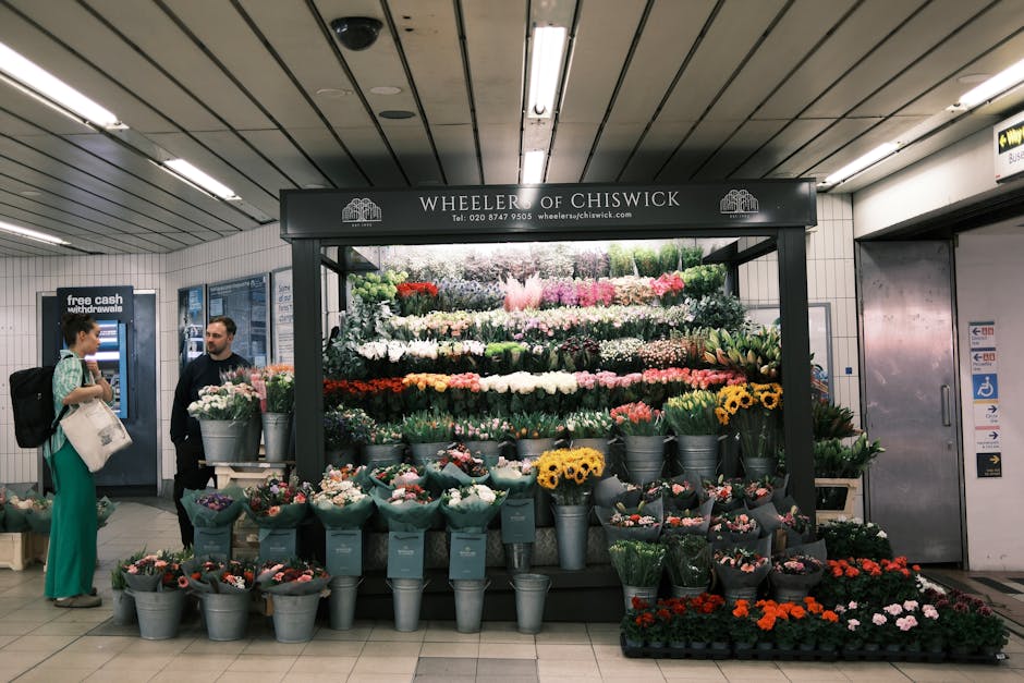 A flower stall named Wheeler of Chiswick located in an underground station, featuring an arrangement of fresh, vibrant flowers including white, pink, red, yellow, and purple blooms. The flowers are di